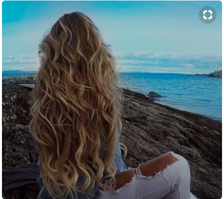 Girl with long, blonde waves sitting by the oceans.